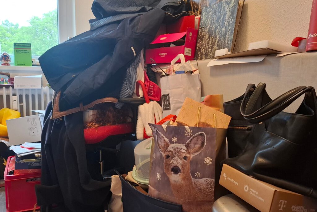 Vorher: Auf dem Boden türmen sich Kleidungsstücke, Kisten und Taschen. Blick ins Arbeitszimmer vor dem Ordnungscoaching: Auf dem Boden türmen sich Kleidungsstücke, Kisten und Taschen.